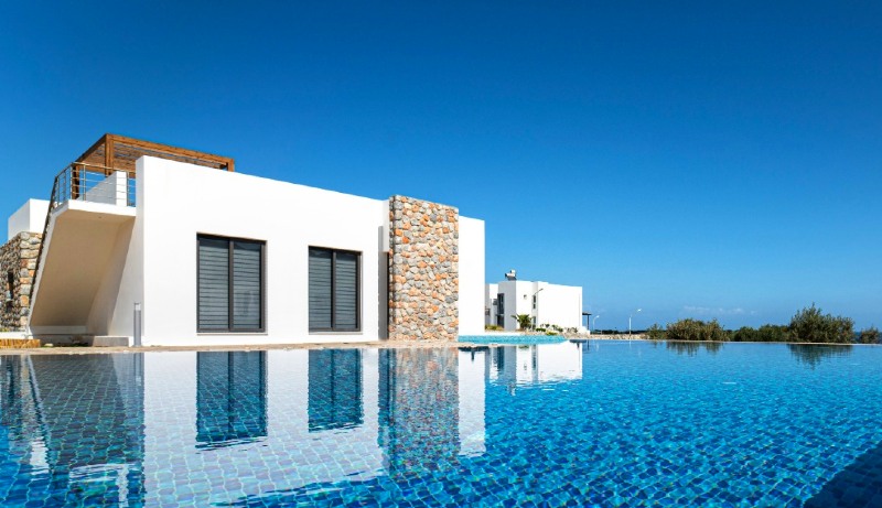 Villa beliggende rett bak et svømmebasseng med knallblåe fliser i. Høy og blå himmel over den hvite sten vill. a