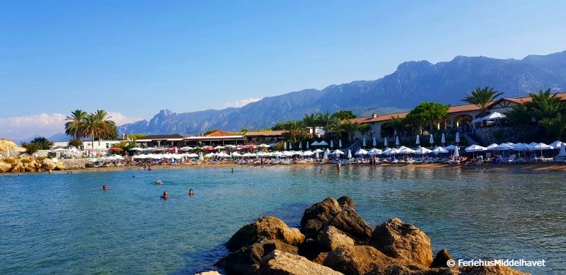 Denizkizi strandklubb med havet foran stranden med solsenger, kafeen bak og helt bakerst fjellryggen Besparmak.